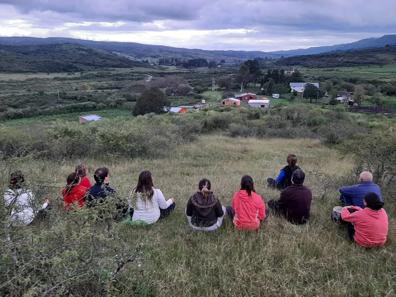 Personas sentadas frente a un paisaje de colinas y construcciones rurales.